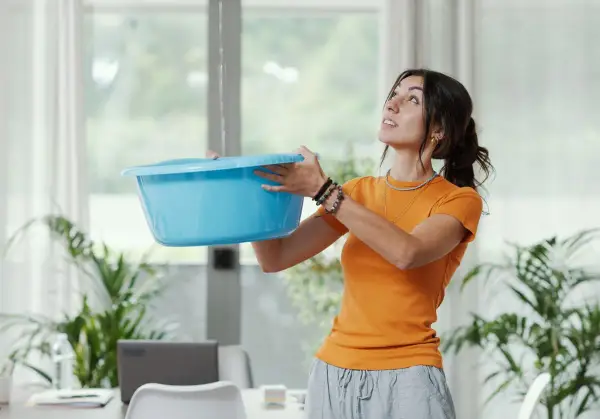 Une femme tente de contenir une fuite d'eau avec un bac bleu, visiblement exaspérée par le dégât des eaux voisin récalcitrant.
